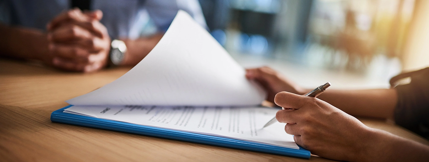 Two people sitting at a table, one with their hands folded and one is reviewing paperwork on a blue folder and is holding a pen in her left hand